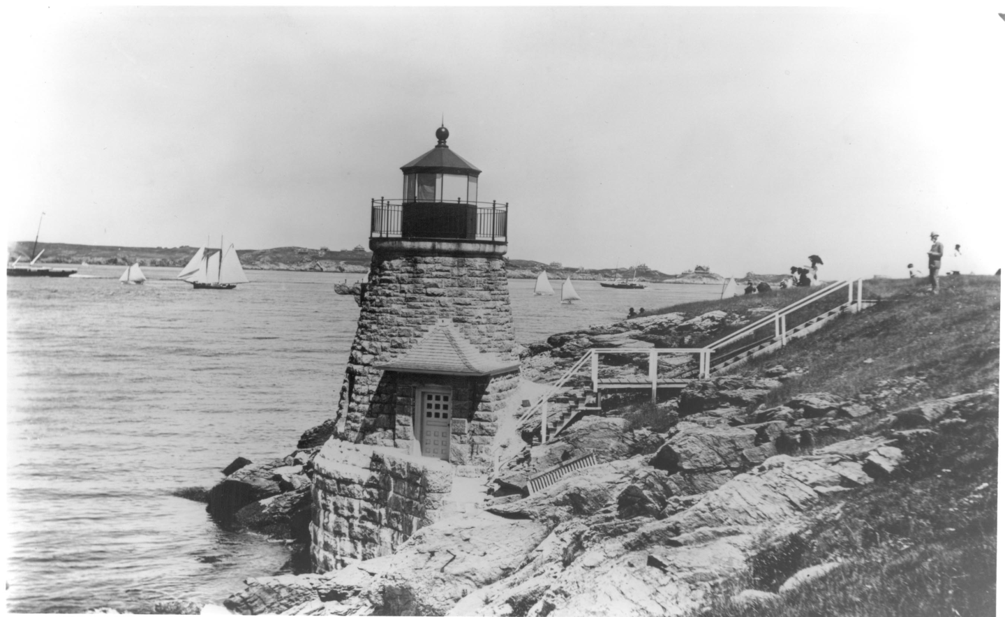 Lighthouse on rocky shore with sailboats in the distance, black and white photograph.