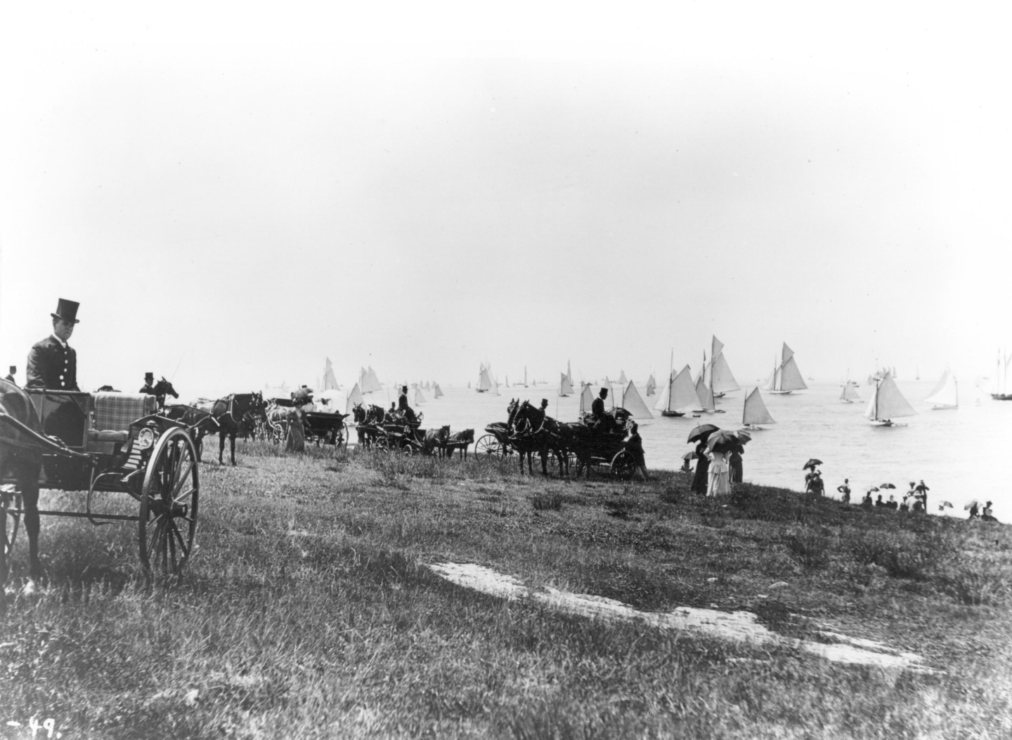 A vintage photo of horse carriages on a grassy shoreline with sailboats in the water.
