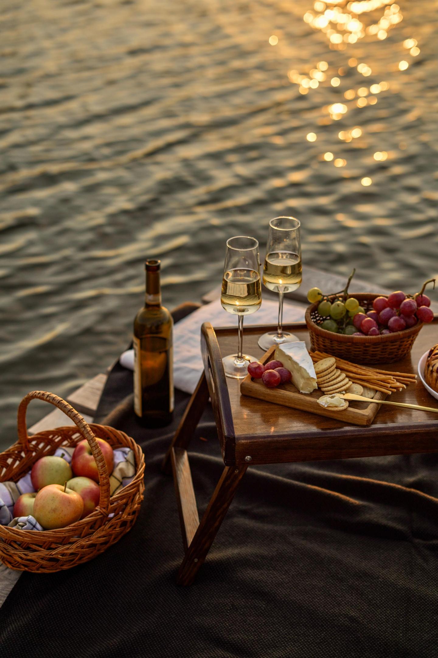 Picnic by the water with wine, cheese, grapes, and apples. Warm sunset lighting.