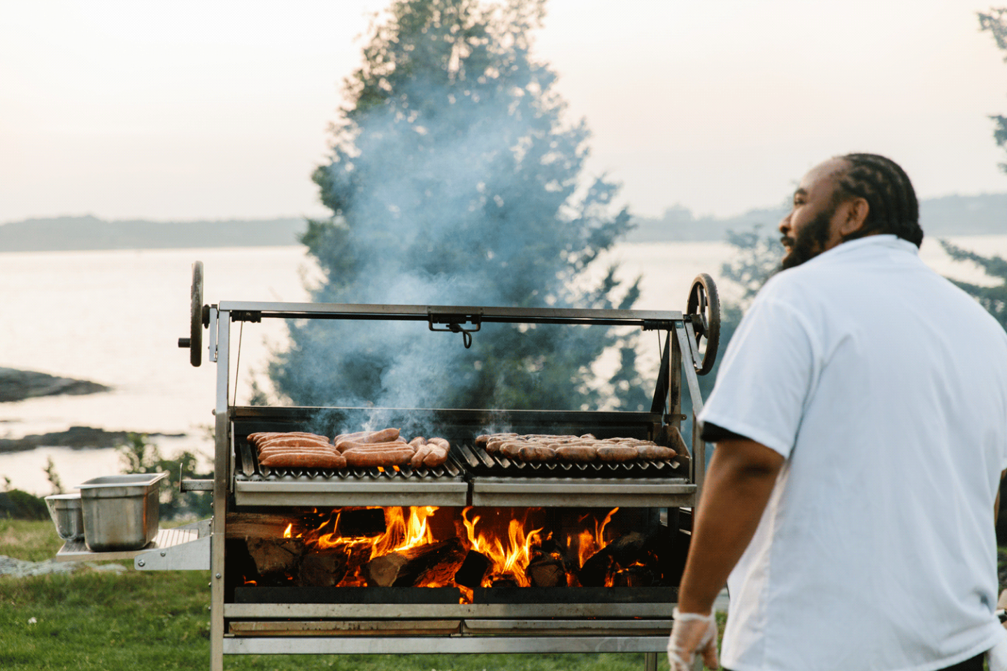 Man grilling sausages outdoors near a lake, with smoke and fire visible.