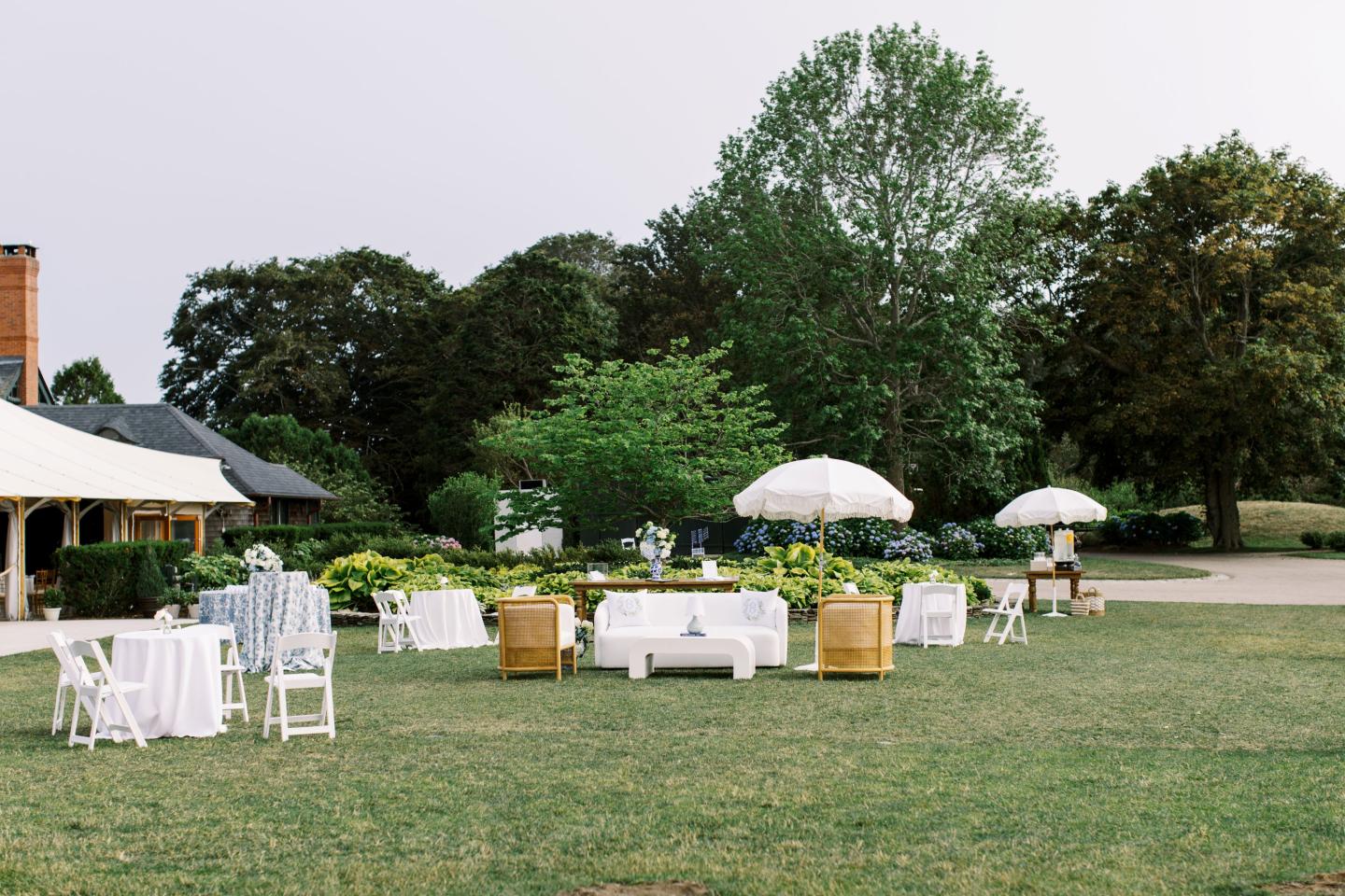 Outdoor event setup with white tables, chairs, and umbrellas on a lawn.