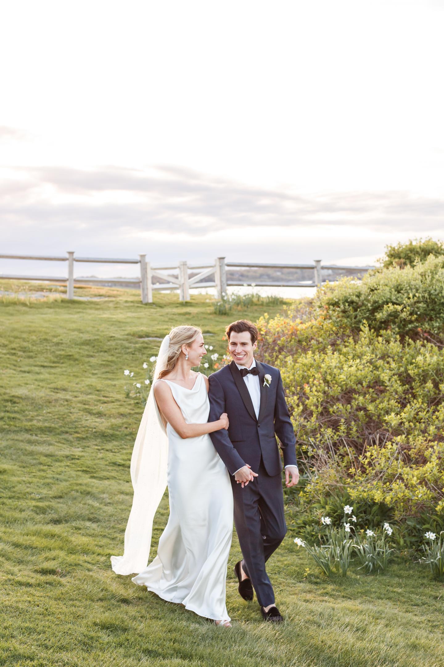 Bride and groom walking on a grassy field, holding hands, with fence in background.