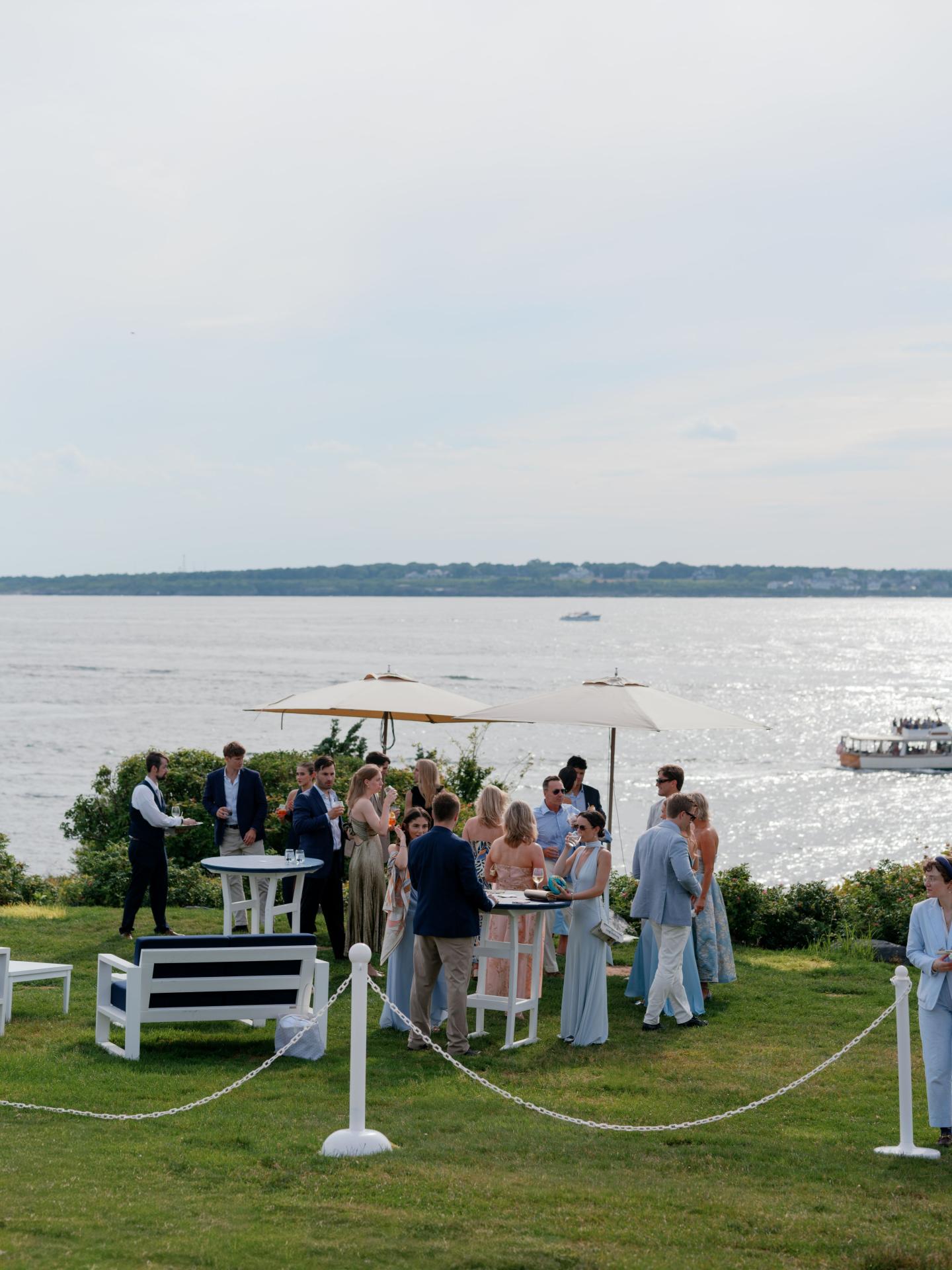 Outdoor gathering by a riverside with people mingling and boats in the background.