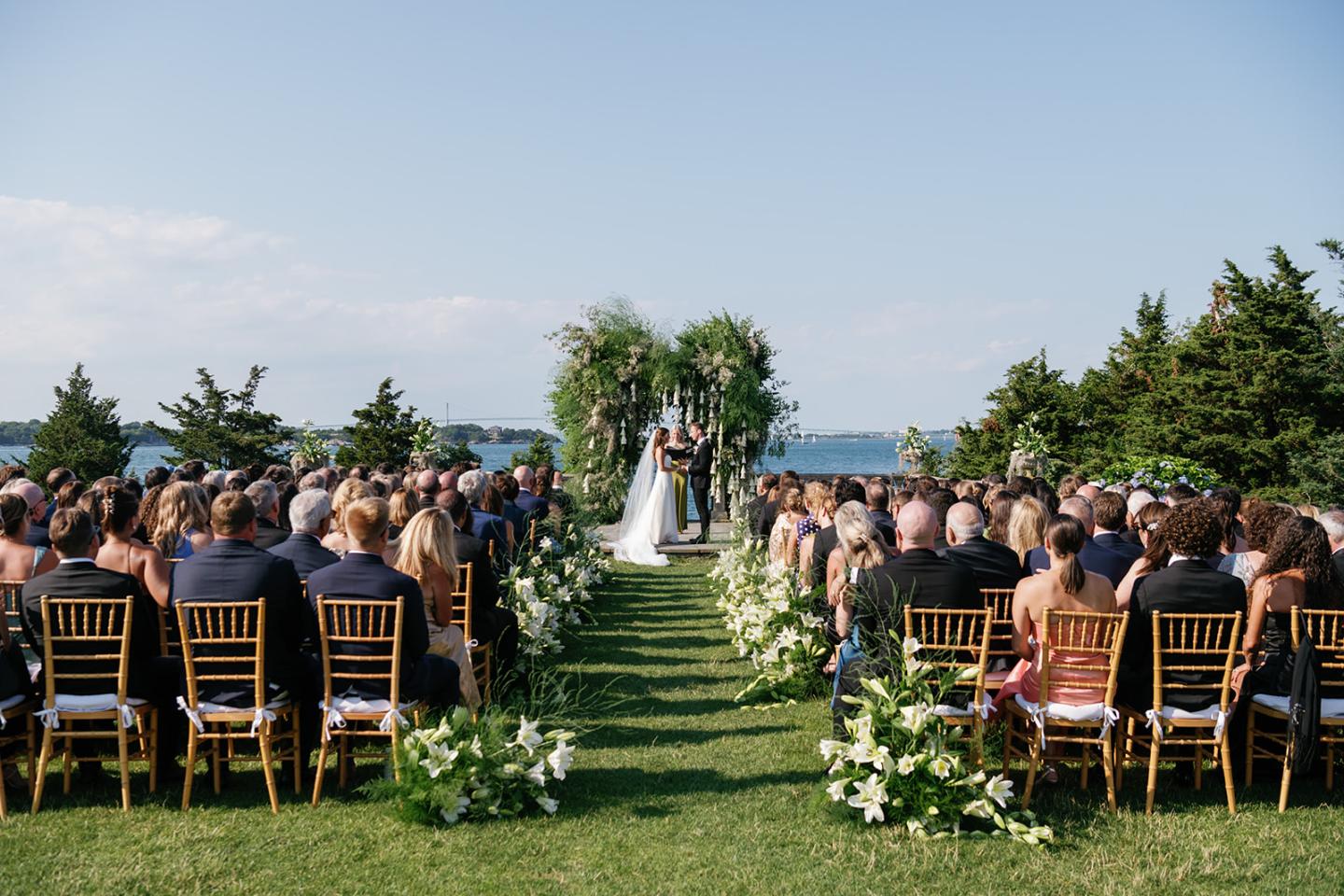 Outdoor wedding ceremony with a couple under an arch, guests seated on both sides.