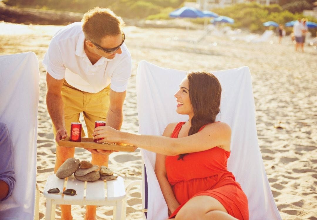 Woman in red shirt in white chair being served drink from man in white shirt on beach at Castle Hill Inn in Newport, RI