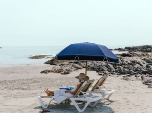Woman on white chair with blue umbrella on rocky beach overlooking ocean at Castle Hill Inn in Newport, RI
