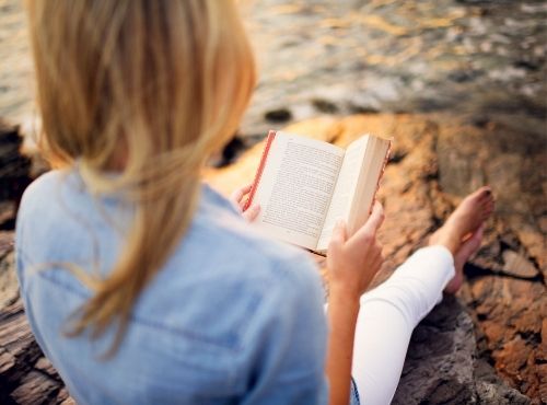 Woman in blue shirt reading a book on rock in front of ocean at Castle Hill Inn in Newport, RI