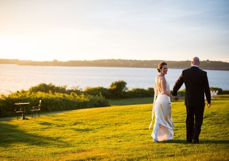 Woman and man walking hand in hand in wedding attire near ocean on top of green grass at sunset at Castle Hill Inn in Newport, RI
