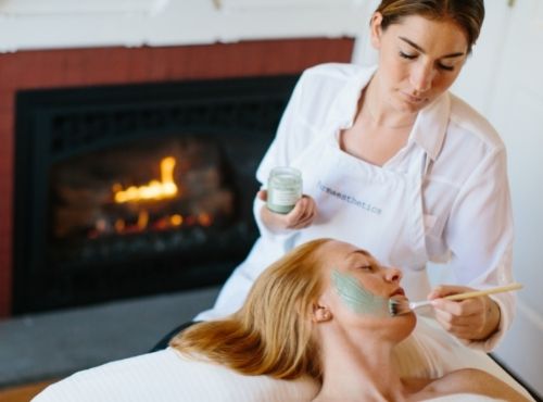 Woman in apron painting facial scrub on to woman's face on table in front of massage oils on top of table at Castle Hill Inn in Newport, RI