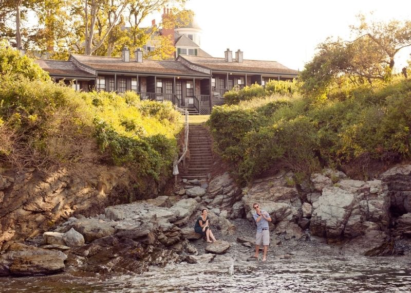 Two people skipping stones on rocky beach in front of house and shrubbery near ocean at Castle Hill Inn in Newport, RI