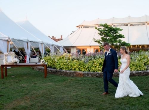 Man and woman in wedding attire holding hands near shrubbery and white tents at Castle Hill Inn in Newport, RI