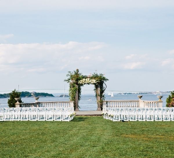Floral archway in front of ocean near stone pillars in front of white chairs on green grass at Castle Hill Inn in Newport, RI